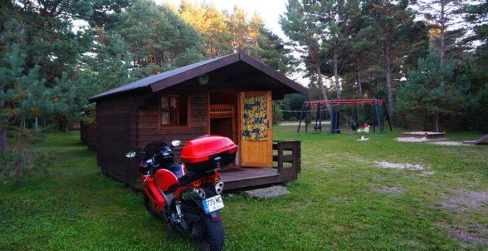 Raudonas motociklas stovi ant žolės šalia medinio namelio.